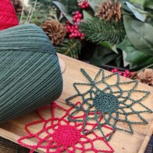 Close up of two blocked crochet snowflake ornaments in red and dark green thread displayed on a wooden serving board with a dark green thread ball holiday greenery pinecones and red berries — Winter Glow Snowflake Ornament pattern by Mistie Bush of American Crochet
