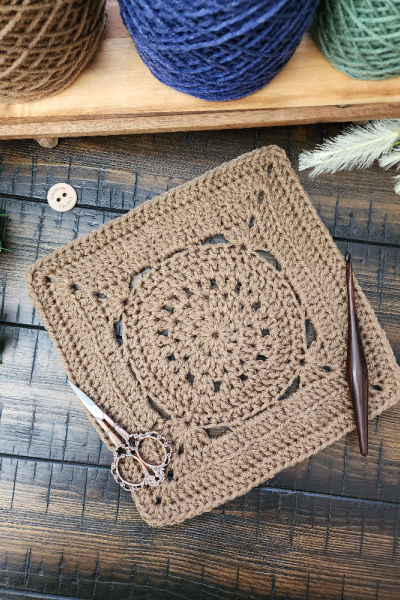 The Pilot Square crochet pattern displayed on a rustic wooden surface with a crochet hook, vintage scissors, and greenery, showcasing its intricate stitch design.
