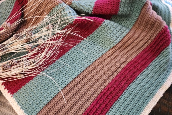 Handmade crochet lapghan featuring horizontal stripes in earthy tones of green, burgundy, and brown, displayed on a wooden surface with a rustic bundle of dried wheat and pinecones resting on top. The textured stitches and cozy palette evoke a sense of warmth and seasonal comfort.