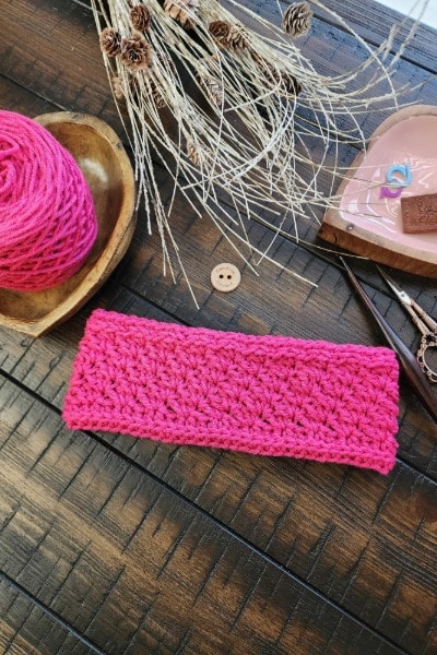 Flat lay of a pink crocheted earwarmer with textured stitches, surrounded by yarn, scissors, stitch markers, and a handmade crochet tag.