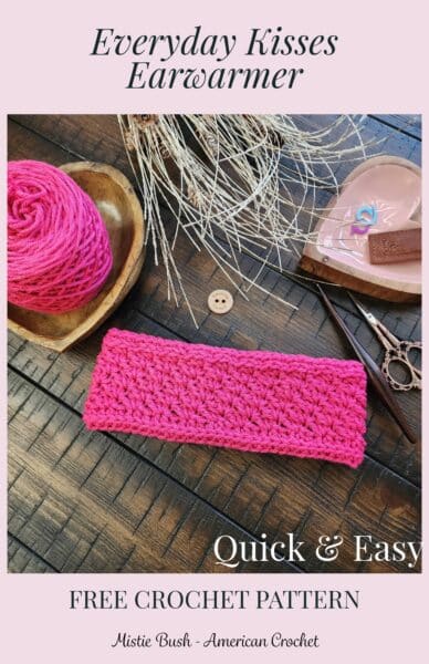 Flat lay of the pink Everyday Kisses Earwarmer on a wooden surface, surrounded by matching yarn in a wooden bowl, ornate scissors, a crochet hook, a handmade wooden button, and stitch markers in a heart‑shaped dish. Text on the image reads ‘Everyday Kisses Earwarmer – Quick & Easy – Free Crochet Pattern by Mistie Bush, American Crochet.