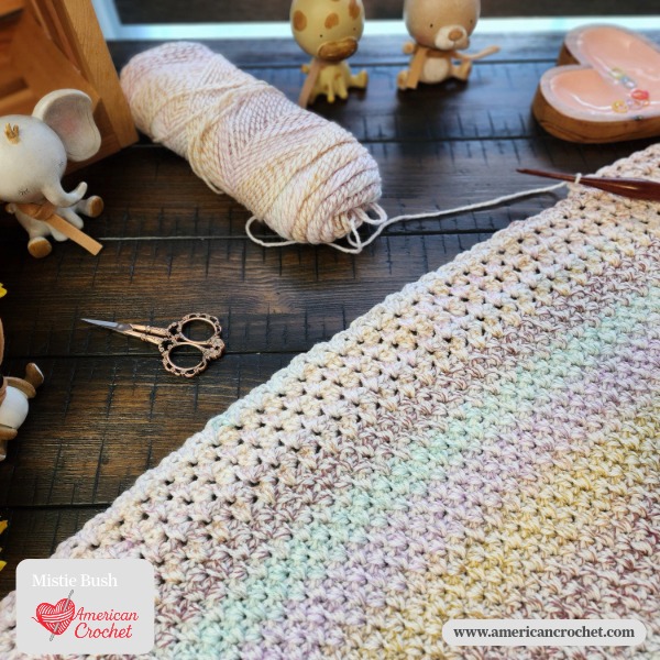 A work-in-progress C2C baby blanket in soft rainbow pastel variegated yarn laid out on a dark wood surface, surrounded by baby animal figurines, ornate scissors, a crochet hook, and a skein of yarn. Mistie Bush / American Crochet branding in the lower left corner.