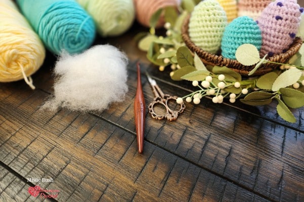 Styled flat lay on a dark wood surface featuring three finished crocheted amigurumi eggs displayed in a woven basket surrounded by eucalyptus stems and white berry sprigs. The eggs show three variations — lavender with purple French knot speckles, blush pink with cream stripes worked in seed stitch, and solid teal. In the upper left three yarn cakes in blush pink, mint green, and soft lavender are arranged alongside a dark wooden crochet hook and decorative scissors. Speckled Nest Eggs Set — Week 3 of the Stitch the Season: Spring Awakening CAL by Mistie Bush of American Crochet.