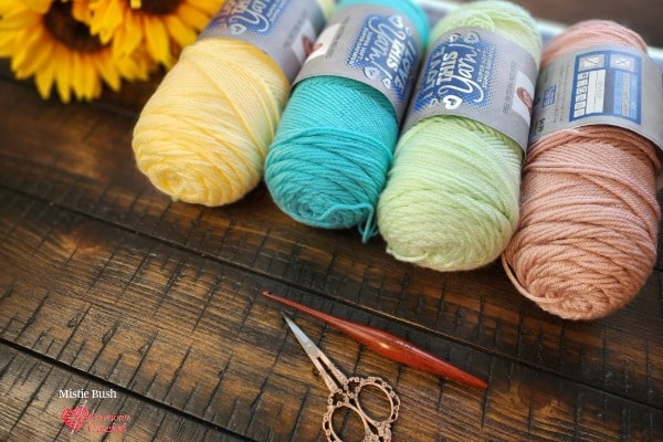 Flat lay on a dark wood surface featuring four yarn cakes in spring pastel colors - Buttercup yellow, Turquoise, Menthe mint green, and Rosy Cheeks blush pink — lined up in a row with a sunflower visible in the upper left corner. A dark wooden crochet hook and decorative scissors rest in the foreground. Yarn selection for the Speckled Nest Eggs Set — Week 3 of the Stitch the Season: Spring Awakening CAL by Mistie Bush of American Crochet.