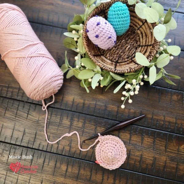 Overhead flat lay of a spring crochet work in progress on a dark wood surface. A blush pink worsted weight yarn cake sits on the left with a partially completed crocheted egg attached to a dark wooden crochet hook. In the upper right a woven basket holds two finished crocheted eggs — one lavender with purple French knot speckles and one teal — surrounded by eucalyptus and white berry stems. Speckled Nest Eggs Set — Week 3 of the Stitch the Season: Spring Awakening CAL by Mistie Bush of American Crochet.