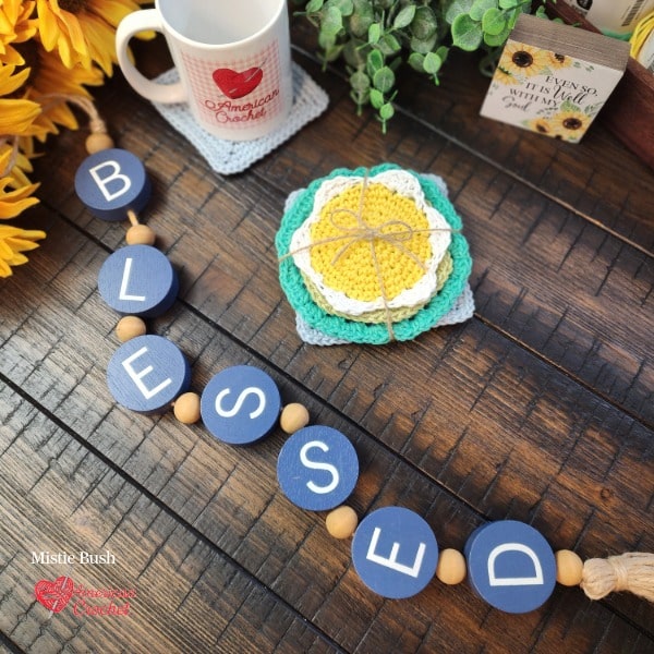 Styled flat lay on a dark wood surface featuring a set of crocheted coasters tied together with natural twine as a gift set. The coaster stack includes a yellow flower coaster, mint green coaster, and gray square coaster arranged together. In the upper left an American Crochet branded white mug sits on a finished square coaster alongside bright yellow sunflowers and lush green eucalyptus. A small decorative sign reading "Even So It Is Well With My Soul" is visible in the upper right corner. A navy blue wooden bead garland spelling "BLESSED" runs diagonally across the foreground. Spring Coaster Set gift styling by Mistie Bush of American Crochet.