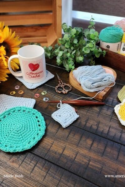 Styled flat lay on a dark wood surface showing the complete Spring Coaster Set collection in progress. Four coaster designs are displayed — a large mint green round coaster with lace border detail, a gray square coaster, a small gray square coaster in progress, and two flower and hexagon coasters in yellow and sage green with white borders. An American Crochet branded white mug sits on a finished square coaster in the upper left alongside bright yellow sunflowers. A wooden heart shaped yarn bowl holds pale blue cotton yarn with a dark wooden crochet hook attached to a coaster in progress. Colorful stitch markers and decorative copper scissors are scattered nearby. A dark wood tray in the upper right holds yarn cakes in mint green, blush pink, sage green, yellow, and white alongside lush eucalyptus. Spring Coaster Set — Week 4 of the Stitch the Season: Spring Awakening CAL by Mistie Bush of American Crochet.