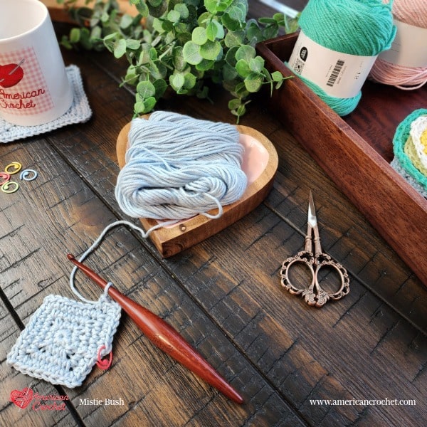Styled flat lay on a dark wood surface featuring a set of crocheted coasters tied together with natural twine as a gift set. The coaster stack includes a yellow flower coaster, mint green coaster, and gray square coaster. In the upper left an American Crochet branded white mug sits on a finished square coaster alongside yellow sunflowers and lush green eucalyptus. A small decorative sign reading "Even So It Is Well With My Soul" is visible in the upper right corner. A navy blue wooden bead garland spelling "BLESSED" runs diagonally across the foreground. Spring Coaster Set gift styling by Mistie Bush of American Crochet.