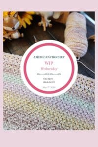 A tall Pinterest image featuring a close-up of a work-in-progress C2C baby blanket in soft rainbow pastel variegated yarn on a dark wood surface. A skein of yarn, ornate scissors, yellow sunflowers, and a wooden fox figurine are visible in the background. A white circle badge in the center reads "American Crochet — WIP Wednesday — One More Skein to Go — May 27, 2026." A soft pink border frames the image.