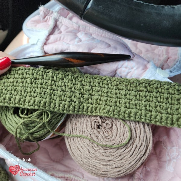 Golden Linen Wrap work in progress showing olive green linen stitch crochet fabric on a decorative hook resting on a skein of taupe yarn inside a project bag — Stitch the Season Golden Hour CAL WIP by Mistie Bush of American Crochet