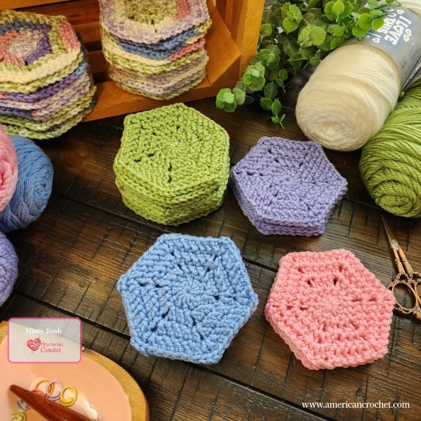 Individual crocheted hexagons in solid green, lavender, blue, and pink arranged on a dark wood surface, with stacks of variegated spring pastel hexagons in the background. Skeins of cream, blue, and green yarn, ornate scissors, greenery, and a wood slice dish with a crochet hook are styled around the hexagons. Mistie Bush / American Crochet branding in the lower left corner.