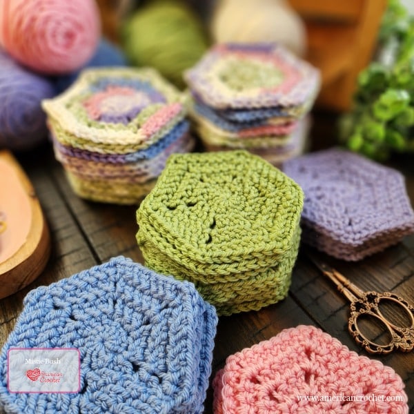 Stacks of crocheted hexagons in spring pastel colors including blue, pink, lavender, and green arranged on a wooden surface with yarn balls and scissors in the background — Petals of Hope Baby Blanket WIP by Mistie Bush of American Crochet.