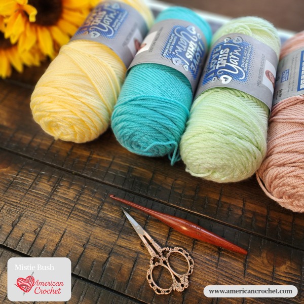 Three skeins of yarn in soft yellow, teal, and mint green displayed with a wooden crochet hook and ornate scissors on a dark wood surface — Mistie Bush, American Crochet.