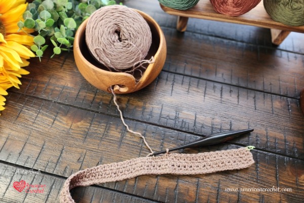 A crochet shawl in progress worked in soft taupe cotton, with the yarn cake in a wooden bowl and a black crochet hook, styled on a dark wood table with a sunflower and autumn-toned yarn — American Crochet.