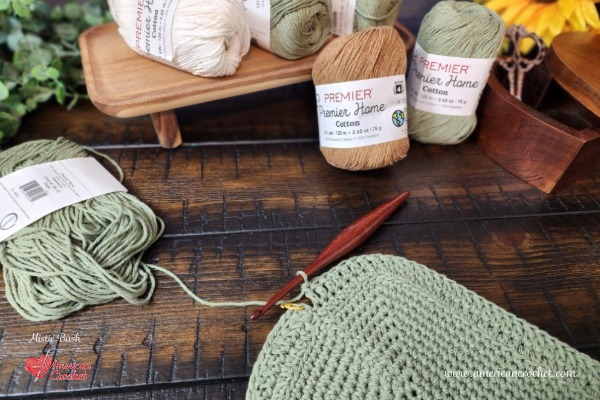 In-progress sage green crochet dishcloth on a wooden hook with Premier Home Cotton yarn skeins and a sunflower — American Crochet