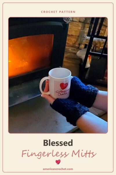 Blessed Fingerless Mitts crochet pattern — navy blue crochet fingerless mitts holding an American Crochet mug by a fire.