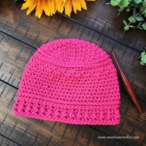 The Reba Beanie crochet hat in pink on a wooden table with sunflowers and a crochet hook